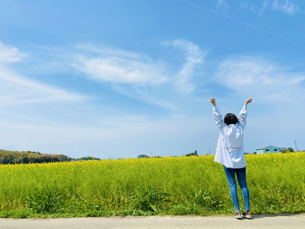開けた菜の花畑と、遠く広がる青空にむかって伸びをする女性の写真。自分のペースでやっていこうと、前向きな気持ちになっていただけるようであれば幸いです。