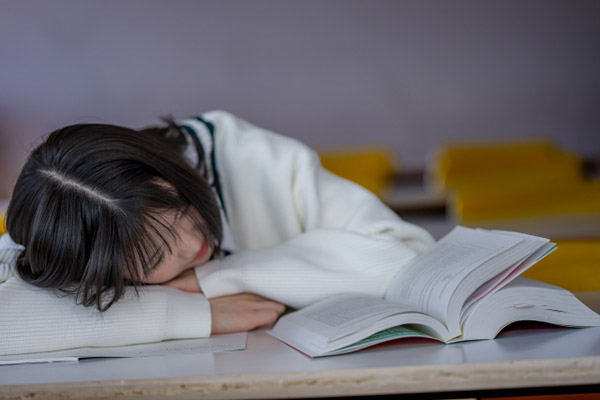 参考書を前に、力なく臥せっている女性の写真。学習のスピードや量についていけそうになく、悩んでいるイメージ。